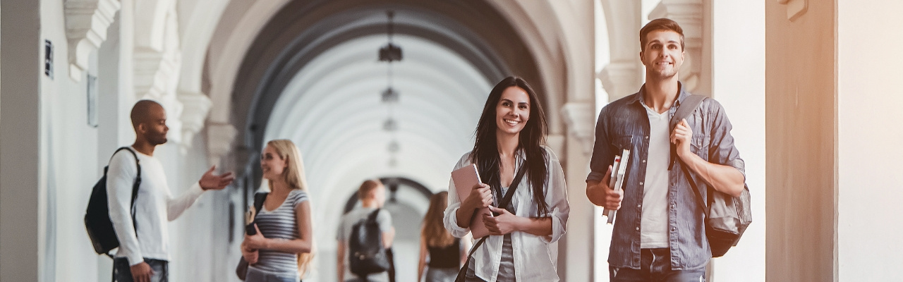 Due ragazzi camminano sotto alle logge universitarie con in mano quaderni e accessori per lo studio. Sullo sfondo altri studenti chiacchierano