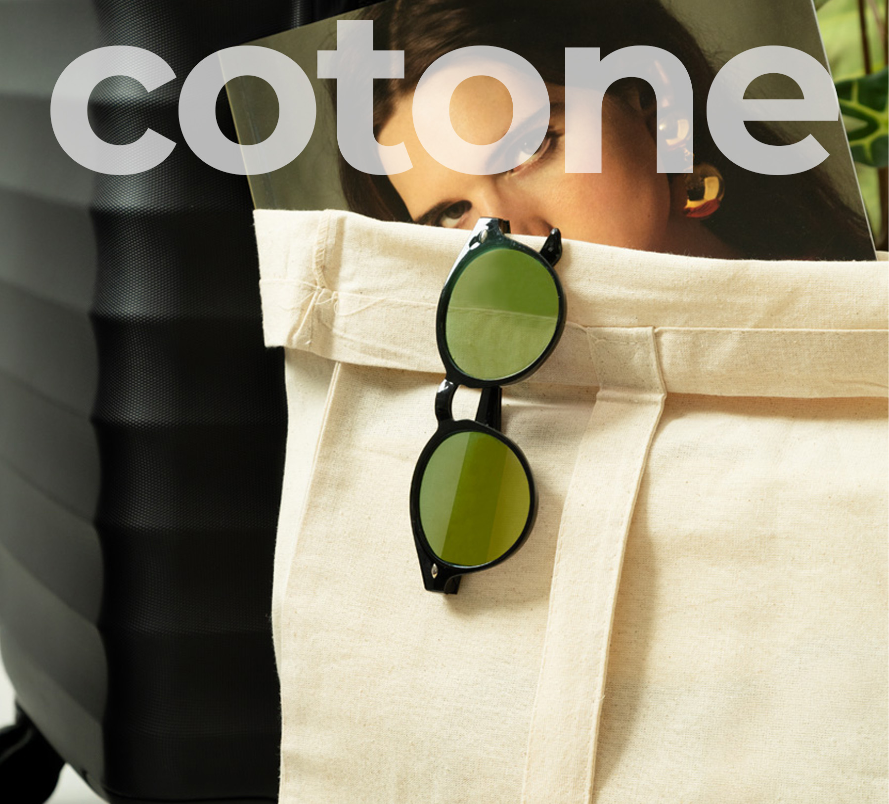 Borsa shopper in cotone naturale beige, con appesi occhiali neri e sullo sfondo trolley nero