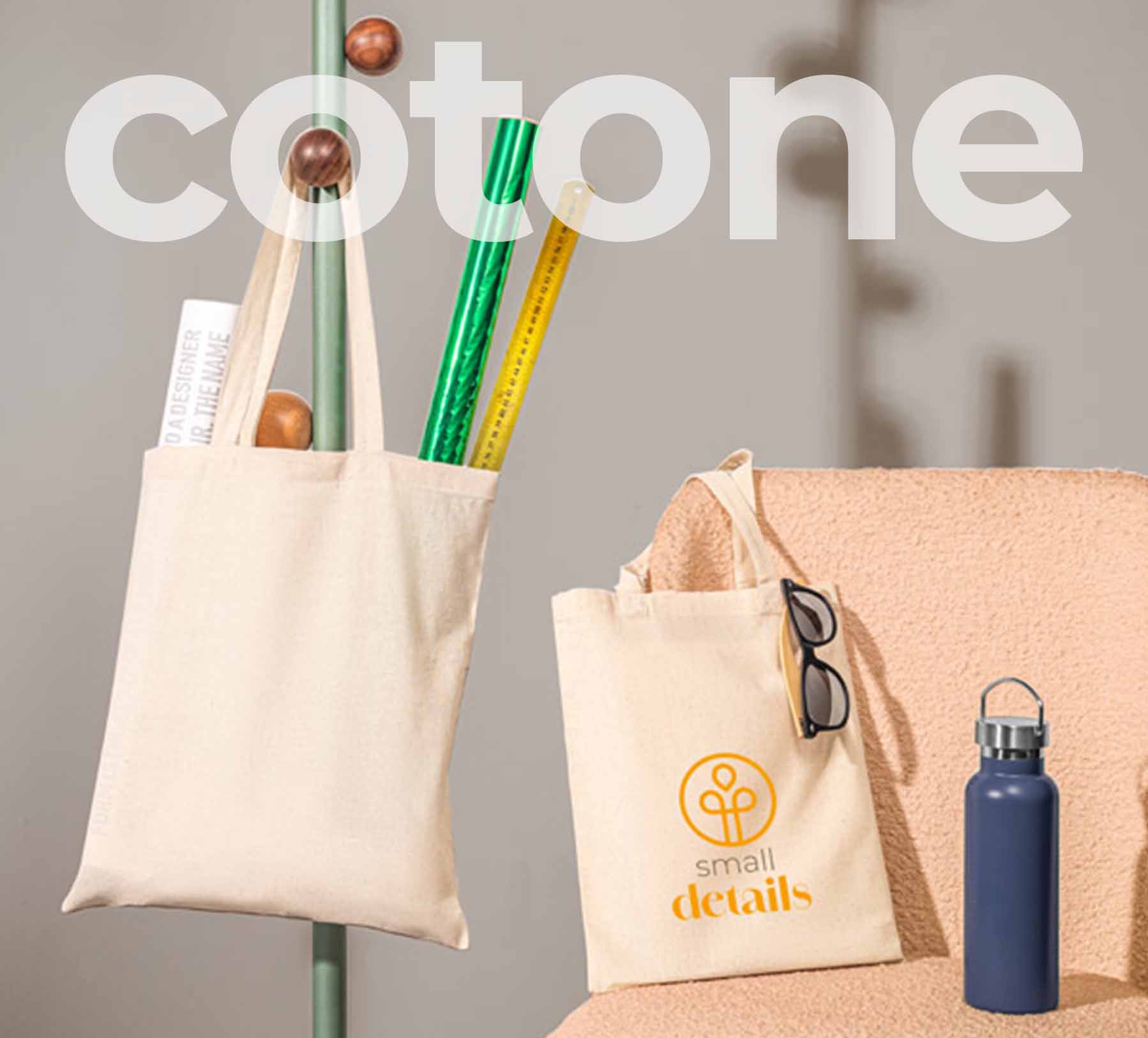 Foto do una shopper in cotone appesa a un appendi abiti con accanto una sedia di tessuto rosa su cui sono appoggiate una borraccia blu e un shopper in cotone con stampa arancione.