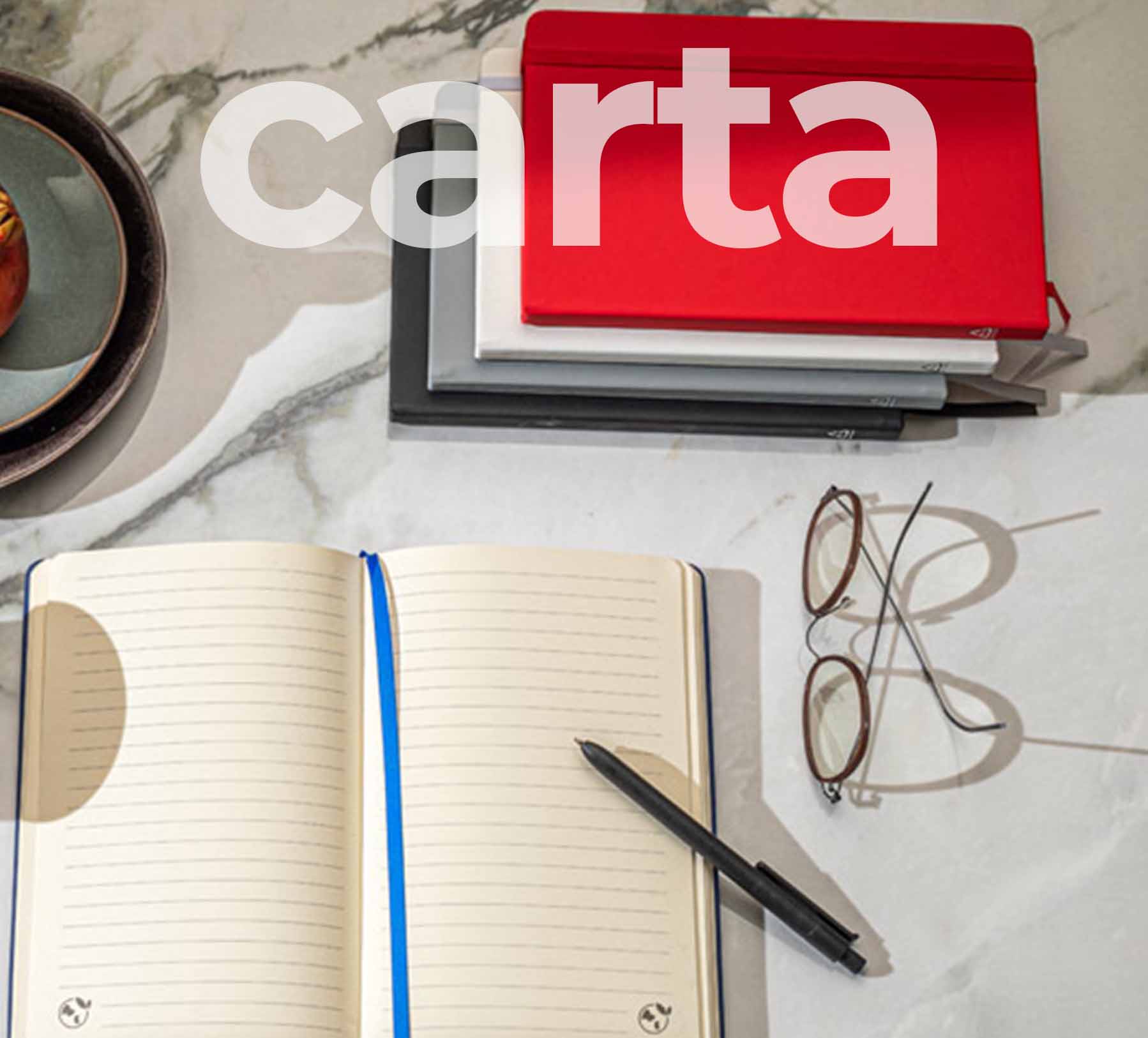 Foto di un block notes con fogli a righe aperto, appoggiato su un tavolo di marmo. Altri block notes di vari colori sono impilati sul tavolo con affianco un paio di occhiali da vista.