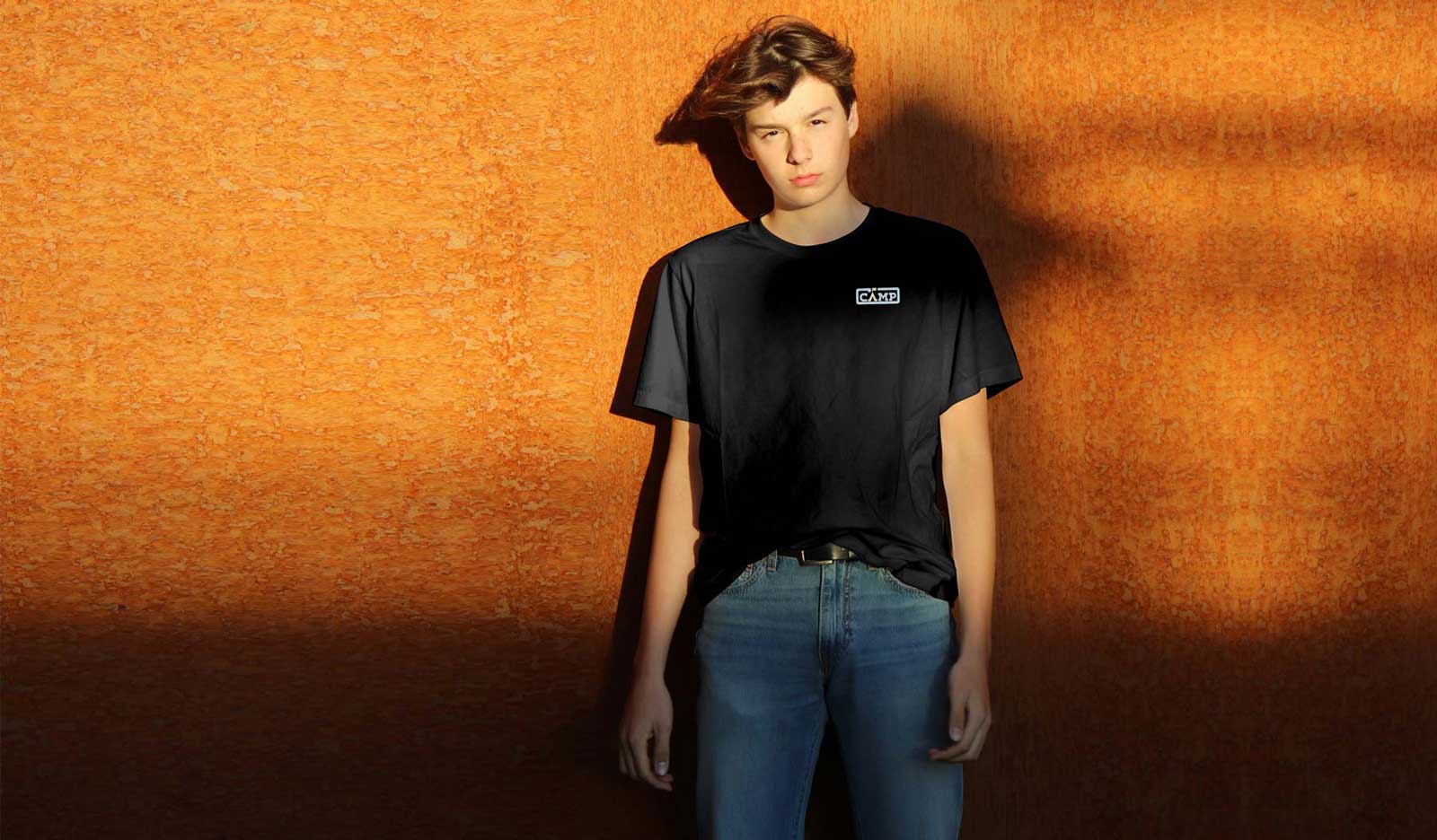 ragazzo in piedi appoggiato a un muro arancione con t-shirt nera con logo colori pastello sul lato cuore e jeans