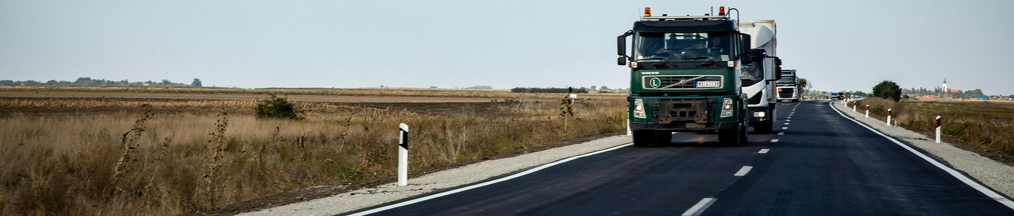 strada con camion che la percorrono e sui lati prati verdi e gialli