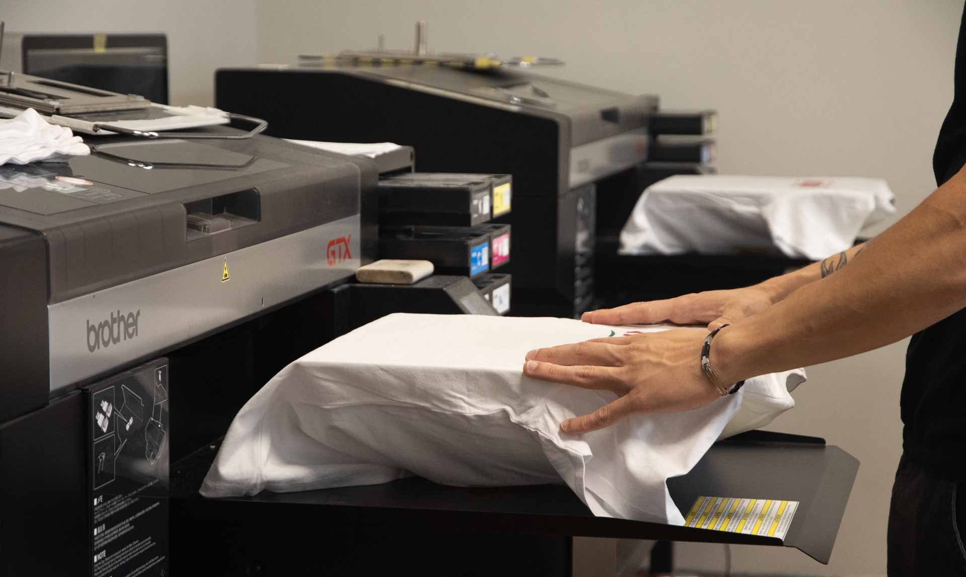 Stampante per tessuti. Sulla destra mani che inseriscono nella dima della stampante una t-shirt bianca.